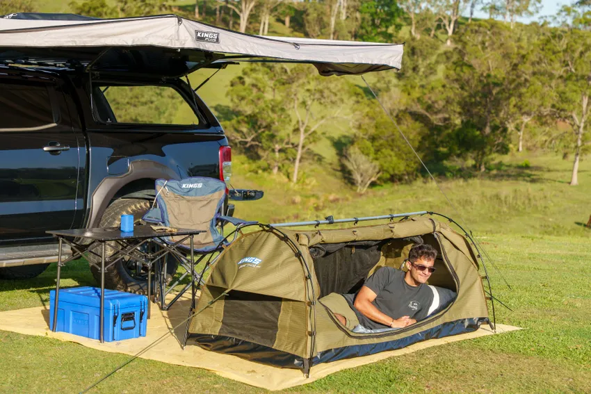 A man relaxing inside an Adventure Kings Big Daddy Deluxe Swag tent at a scenic campsite with a 4x4 vehicle and awning setup.