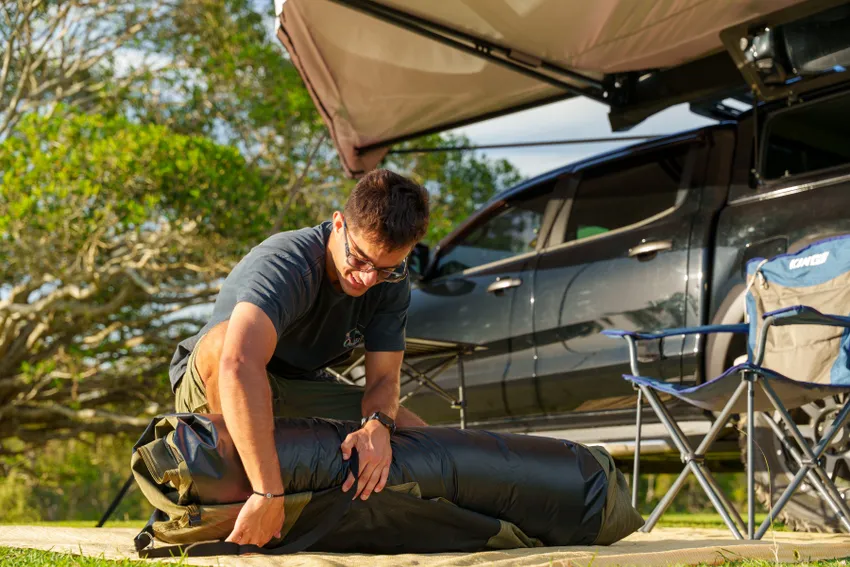 A man packing up an Adventure Kings camping swag on a sunny day, with a 4x4 vehicle and camping awning in the background.